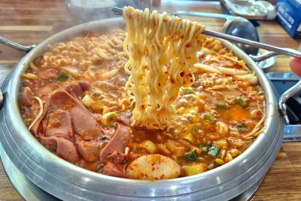Ramyeon noodles cooking in a bubbling pot of budae jjigae with ham and sausage.