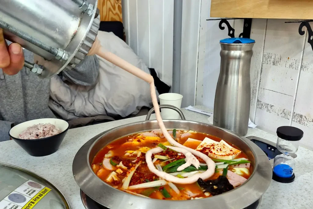 Fresh house-made ham being added to budae jjigae with a Vulcan cannon-style device at a restaurant in Daejeon.