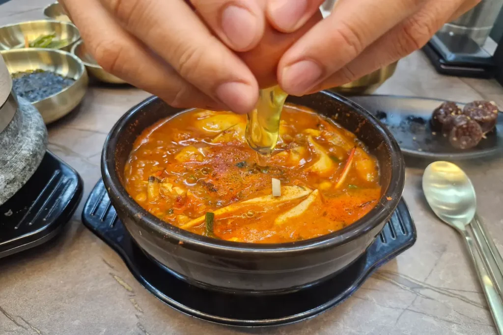 A raw egg being cracked into a bubbling pot of Korean sundubu jjigae.